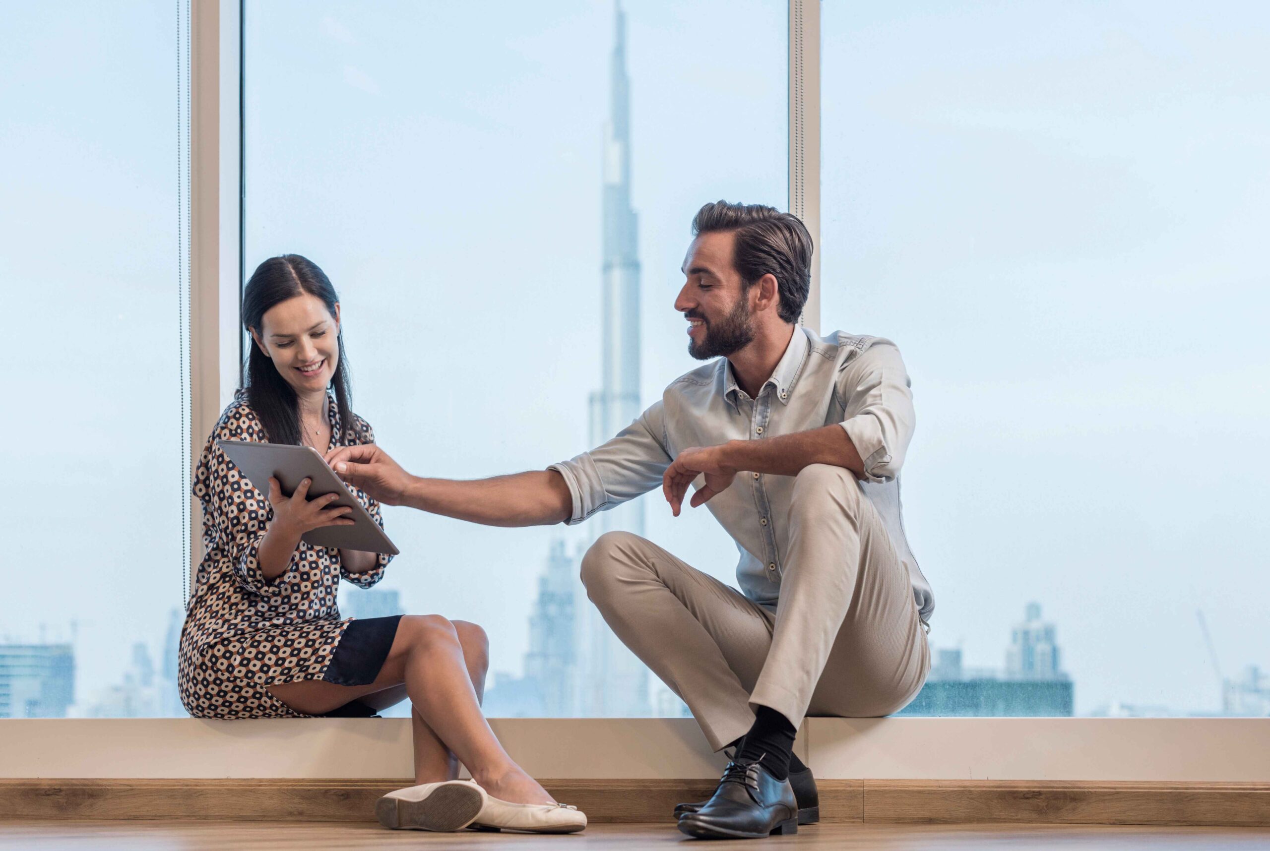 A stylish couple sitting by a large window with the Dubai skyline in the background, smiling and discussing property options on a tablet, symbolizing clarity and confidence after a virtual tour.