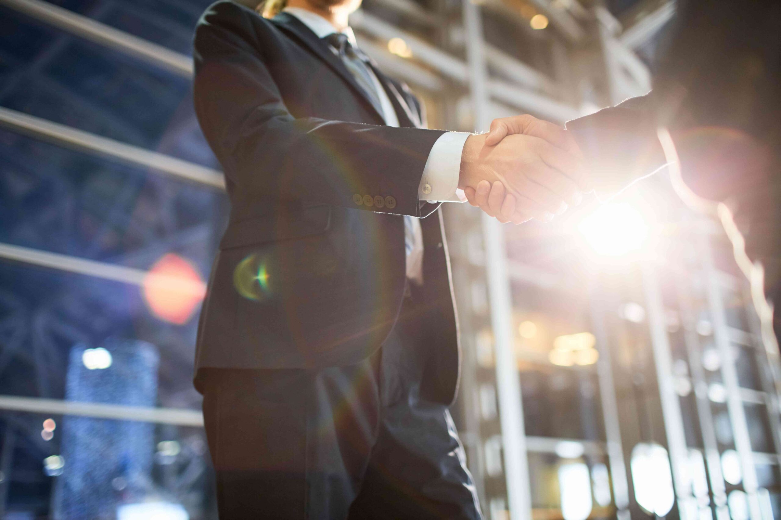 Two business professionals in suits shaking hands in a modern glass building with sunlight flaring behind them, symbolising partnership, approval, and a trusted reseller collaboration with Exclusive Luxe Tours.